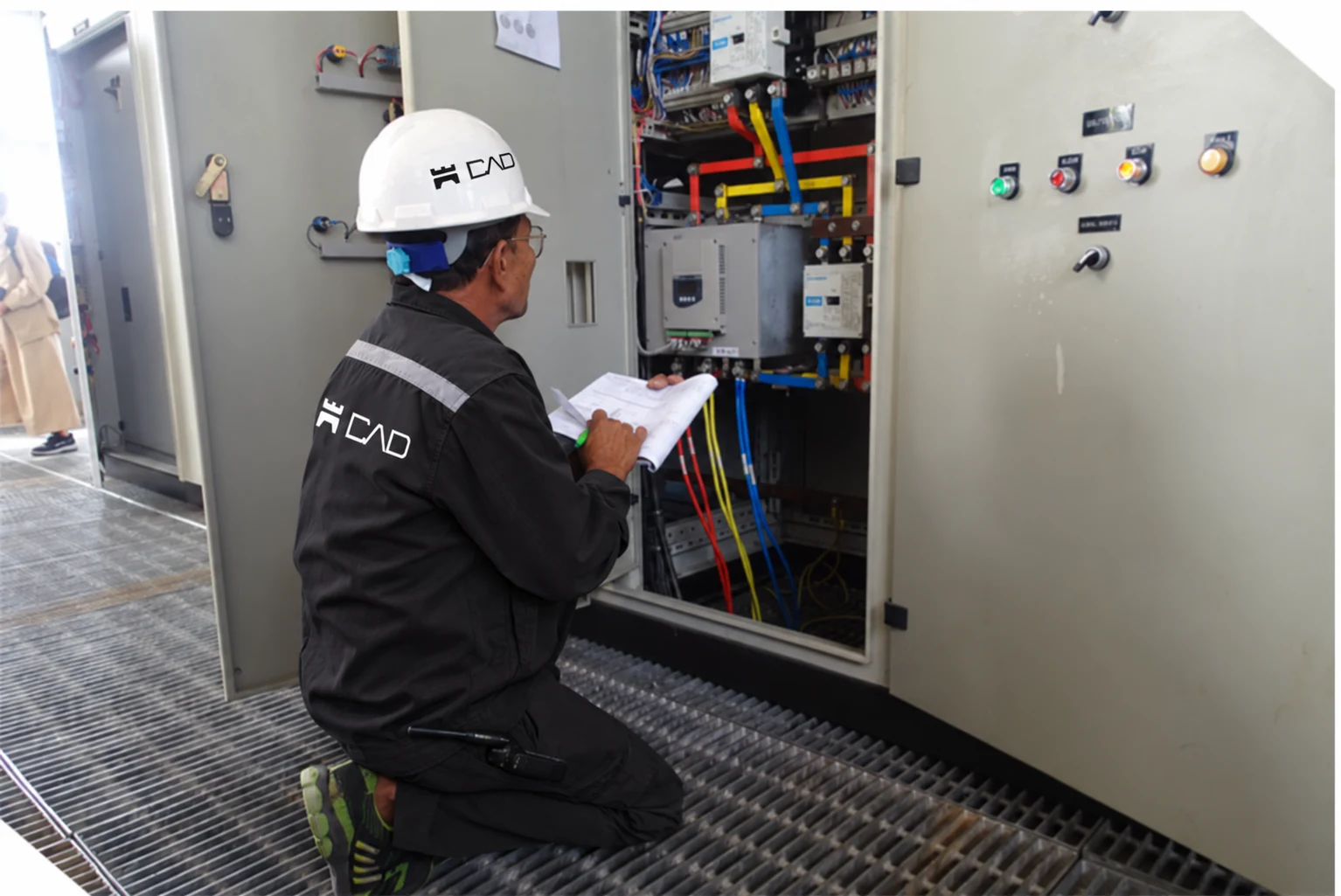 Profissional com uniforme preto e capacete com a marca CAD inspecionando painel elétrico industrial